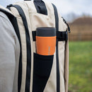 A person carries a beige backpack with a black pocket, which holds the Lifeventure One-Touch Thermal Mug in orange—a stainless steel mug featuring a convenient one-touch lid. The backdrop is grassy with a softly blurred outdoor scene.