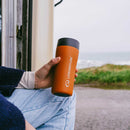 A person in jeans and a dark sweater holds a vibrant orange Lifeventure One-Touch Thermal Mug with its convenient one-touch lid while sitting by an open door, with grass and the sea visible in the background.