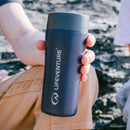 A person holding a Lifeventure One-Touch Thermal Mug in navy blue outdoors, with rocks and another person visible in the background.