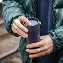A person wearing a green patterned jacket holds the Lifeventure One-Touch Thermal Mug, made of dark stainless steel, with both hands and displays several silver rings on their fingers. The background is blurred, likely outdoors.