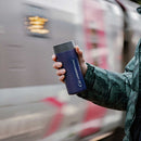 A person in a green jacket holds a blue Lifeventure One-Touch Thermal Mug featuring a convenient One-Touch Lid, while a blurred train passes by in the background.