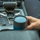 A hand places a Lifeventure One-Touch Thermal Mug, featuring blue and black stainless steel with a one-touch lid, into a car’s cupholder next to the gear shift and control buttons.