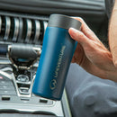 A hand holds a Lifeventure One-Touch Thermal Mug in blue, featuring a convenient one-touch lid, inside a car above the center console and dashboard controls.