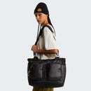 A person wearing a black beanie and white shirt carries The North Face Base Camp Voyager Tote Bag, crafted from recycled durable fabric and featuring two front pockets, over their shoulder while standing against a plain light background.