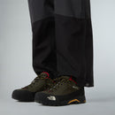 A person is standing on a grey background wearing black outdoor pants and The North Face Men’s Verto GORE-TEX Alpine Shoes in olive green with yellow and red laces and a Vibram outsole.