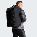 A person in a gray jacket and sunglasses carries a large black Great Outdoors Base Camp Boot Bag on their back, facing left against a plain light background.
