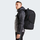 A person in a gray puffer jacket and sunglasses carries the Great Outdoors Base Camp Boot Bag on their back, facing sideways against a plain light background.