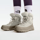 Close-up of a person wearing light gray Womens ThermoBall Leather Waterproof Boots by The North Face, featuring thick soles and faux fur lining, styled with white The North Face socks and dark purple leggings.