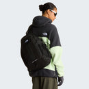 A person wearing sunglasses, a black and light green The North Face fleece jacket, and gloves carries a sleek Hot Shot Sling Bag by The North Face over one shoulder, along with a black The North Face backpack, while facing away from the camera.