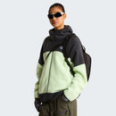 A person wearing black sunglasses, a black and light green fleece jacket, and olive pants with The North Face Hot Shot Sling Bag stands against a plain light background.