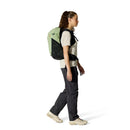 A woman with a ponytail, dressed in a beige shirt, dark pants, and sneakers, walks while carrying the Rab Airox ND22L Day Pack with a ventilated AEROFIT back system, set against a plain white background.