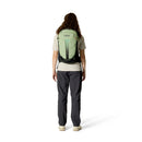 A person with long curly hair, dressed in a light beige shirt and dark gray pants, stands facing away against a white background while carrying the Rab Airox ND22L Day Pack—a women’s hiking backpack featuring a ventilated AEROFIT back system.