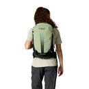 A person with long hair, viewed from behind, wears a light green Rab Airox ND22L Day Pack—a women’s hiking backpack designed with a ventilated AEROFIT back system—as well as outdoor clothing, including a beige shirt and gray pants, set against a plain white background.