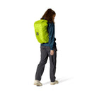 A person in outdoor clothing stands sideways, wearing a bright green backpack cover over a large women’s hiking backpack, such as the Rab Airox ND22L Day Pack with its ventilated back system. The background is plain white.