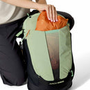 A person is placing an orange bag into the front mesh pocket of a light green and black Rab Airox ND22L Day Pack, a women’s hiking backpack. Only the person’s hand, part of their torso, and legs are visible. The backpack features Rab’s ventilated AEROFIT back system for enhanced comfort on the trail.