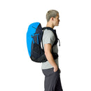 A person with short hair, dressed in a light gray shirt and dark pants, is standing in profile with the Rab Airox 30L Hiking Pack—featuring an AEROFIT back system, and colored blue and black—on their back, isolated on a white background.