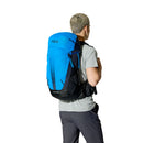 A person dressed in a light gray shirt and dark pants carries the Rab Airox 30L Hiking Pack in blue, featuring black straps. Facing away from the camera against a white background, they highlight the backpack’s ventilated AEROFIT back system, designed by Rab for enhanced comfort during outdoor adventures.