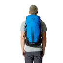 A person with short hair, dressed in a grey t-shirt and dark pants, is facing away from the camera and carrying a Rab Airox 30L Hiking Pack in blue and black, which features the ventilated AEROFIT back system.