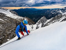 A mountaineer in a blue jacket, blue helmet, and red pants ascends a snowy mountain slope under a dramatic cloudy sky with distant rugged peaks.