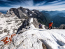 Two climbers in bright gear, equipped with Scarpa Men’s Mont Blanc Pro GTX technical mountaineering boots, ascend a snowy, rocky mountain ridge under a dramatic sky, with rugged peaks and a deep valley stretching into the distance.