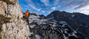 Person climbing a rocky mountain with a scenic view of snow-capped peaks and blue sky.