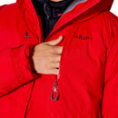 A person in a vibrant red Rab Batura Jacket touches their chest near a zipped pocket, highlighting the jacket’s front features and high collar—perfect for high-altitude expeditions with its premium 800FP European goose down insulation.
