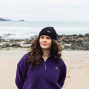 Woman wearing a black beanie and purple fleece jacket on a beach