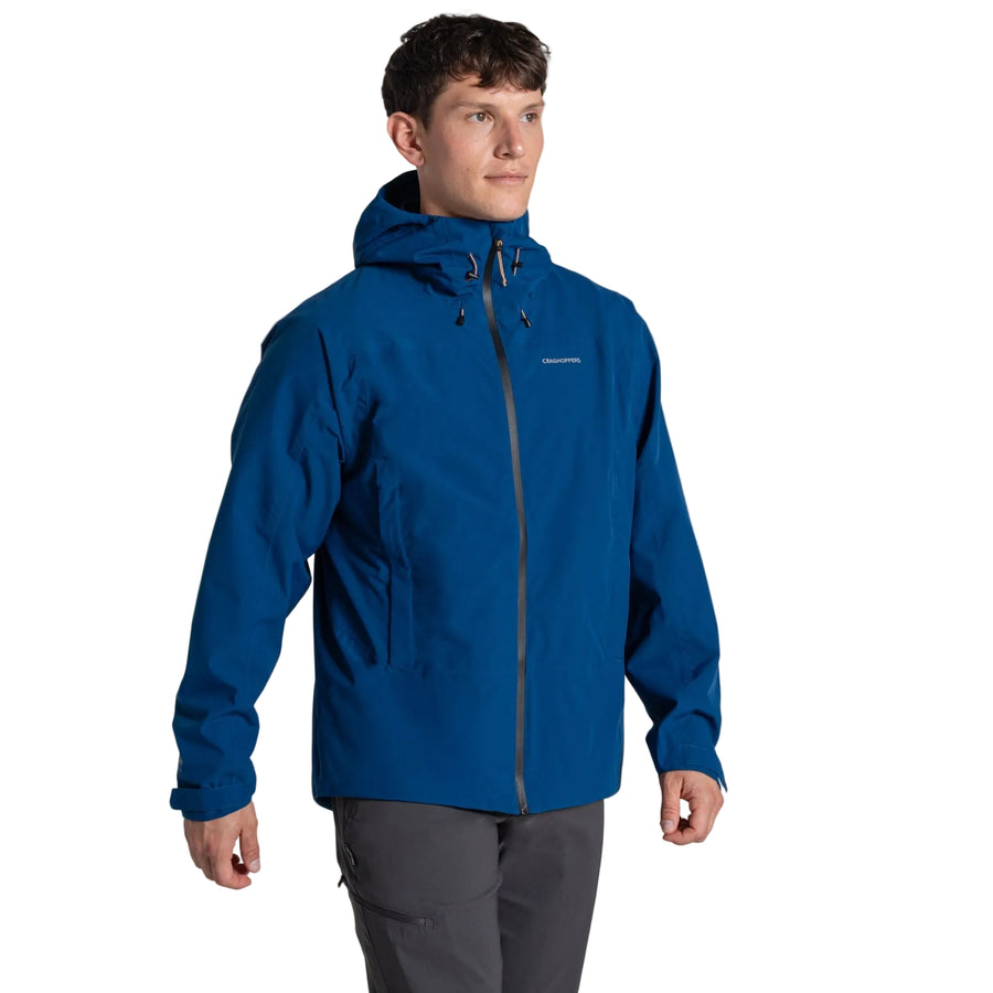 A man stands facing forward against a white background wearing the Craghoppers Mens Ponsa Waterproof Jacket in blue with AquaDry Membrane, paired with gray pants.