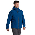 A man stands facing forward against a white background wearing the Craghoppers Mens Ponsa Waterproof Jacket in blue with AquaDry Membrane, paired with gray pants.