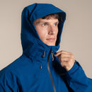 A man wearing the Craghoppers Mens Ponsa Waterproof Jacket in blue looks to the side and zips it up with one hand against a plain beige background.
