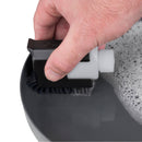 A hand uses a small cleaning brush with an attached bottle from the Sea to Summit Camp Kitchen Clean Up Kit (6-Piece) to scrub a soapy gray surface—possibly a dish or pan—with suds visible on part of the surface.