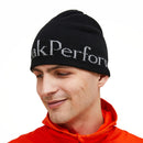 A person wearing a black Peak Performance PP Hat with white text and an orange jacket, looking down and smiling against a white background. The double-layer knit construction of the PP Hat provides warmth and comfort.