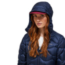A person with long red hair wears the Peak Performance Womens Helium Down Hood Jacket in blue, with the hood up and a red-brimmed cap underneath, looking at the camera against a plain background.