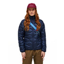 A person with long red hair, wearing a maroon cap, tan pants, a blue crossbody bag, and the Peak Performance Womens Helium Down Hood Jacket in navy blue stands facing forward against a plain white background.