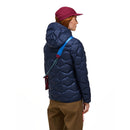 A person is shown from the back against a plain white background, wearing a maroon cap, brown pants, and the Peak Performance Womens Helium Down Hood Jacket—a blue insulated jacket with a windproof, water-repellent shell—along with a blue strap across their shoulder.