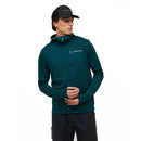 A man wearing the Peak Performance Mens Trail Polartec Power Grid Zip Hooded Fleece in dark green, paired with a black baseball cap. This technical midlayer features a chest logo and is crafted from Polartec fleece. He stands against a plain white background, looking to the side with his hands clasped in front.