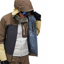 A person wearing the Peak Performance Mens Vislight GORE-TEX C-Knit 3L Shell Jacket in brown, yellow, and gray ski touring style, with the hood up and ski goggles, opens the lightweight waterproof shell to display the interior lining and pocket, revealing a puffy white inner layer. The background is plain white.