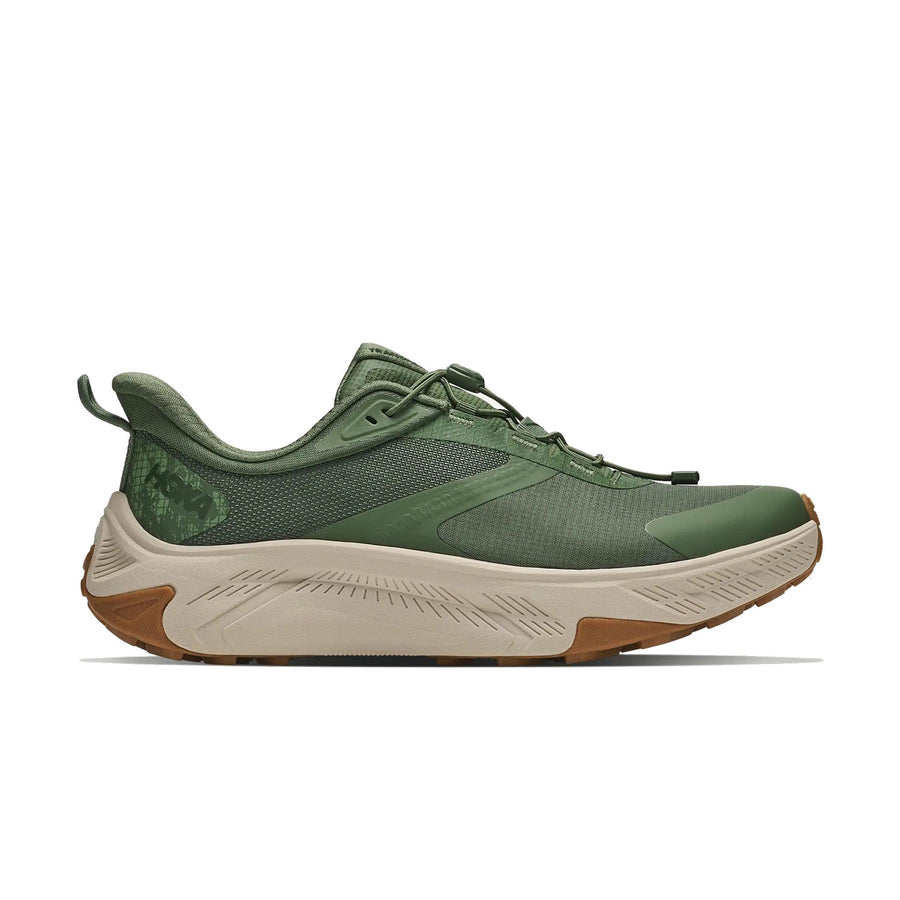 A green Hoka Mens Transport 2 sneaker with a chunky beige sole and refined textured accents is displayed in side profile against a white background, featuring sustainable cushioning engineered for running on both city streets and trails.