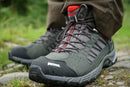 A close-up of a person wearing Meindl Mens Journey Mid GTX boots, featuring the Comfort Sport sole, standing on a rocky path with green grass in the background.