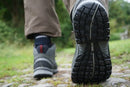 Close-up of a person walking on a stone path, showcasing the tread of the Meindl Mens Journey Mid GTX hiking shoe with Gore-Tex lining. The Meindl Comfort Sport sole is visible. Green grass and trees are blurred in the background.