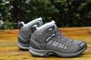 A pair of Meindl Womens Journey Mid GTX Walking Boots in gray with blue and white accents rests on a wooden surface outdoors, set against a backdrop of blurred greenery.