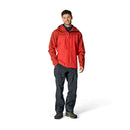 A man stands smiling, wearing the Rab Mens Latok Mountain GORE-TEX Pro Jacket in red, along with dark waterproof pants and hiking shoes, against a plain white background.