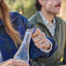 Outdoors amid blurred greenery, one warmly dressed person uses the Leatherman Skeletool® KB Knife to open a glass bottle, while another sits beside them.