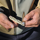 Someone holds and opens a Leatherman Skeletool® KB Knife with a tan handle and 420HC stainless steel blade, while wearing a beige and green jacket.