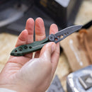 A person holds a Leatherman Skeletool® KB Knife with a green handle and partially open 420HC stainless steel blade featuring cut-out holes, indoors with various blurred objects in the background.