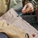 Wearing an outdoor jacket, a person uses the Leatherman Skeletool® KBx Knife to open a crumpled brown paper bag while sitting outside.