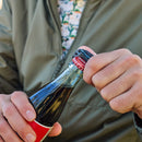 Wearing a green jacket and patterned shirt, someone uses the Leatherman Skeletool® KBx Knife, which has a 420HC stainless steel blade, to open a glass soda bottle.