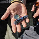 A hand holds a Leatherman Skeletool® KBx Knife and a bottle cap, while the other grips a brown glass bottle. The person, dressed in dark pants and a light jacket, is sitting outdoors.