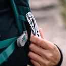 Outdoors, someone clips a Leatherman Skeletool® KBx Knife with a 420HC stainless steel blade onto the strap of a green backpack.