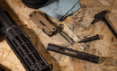 A disassembled firearm and a Leatherman Super Tool® 300M - Molle Black sit on a wooden surface alongside a charging handle and cleaning patches, indicating firearm maintenance or cleaning underway.